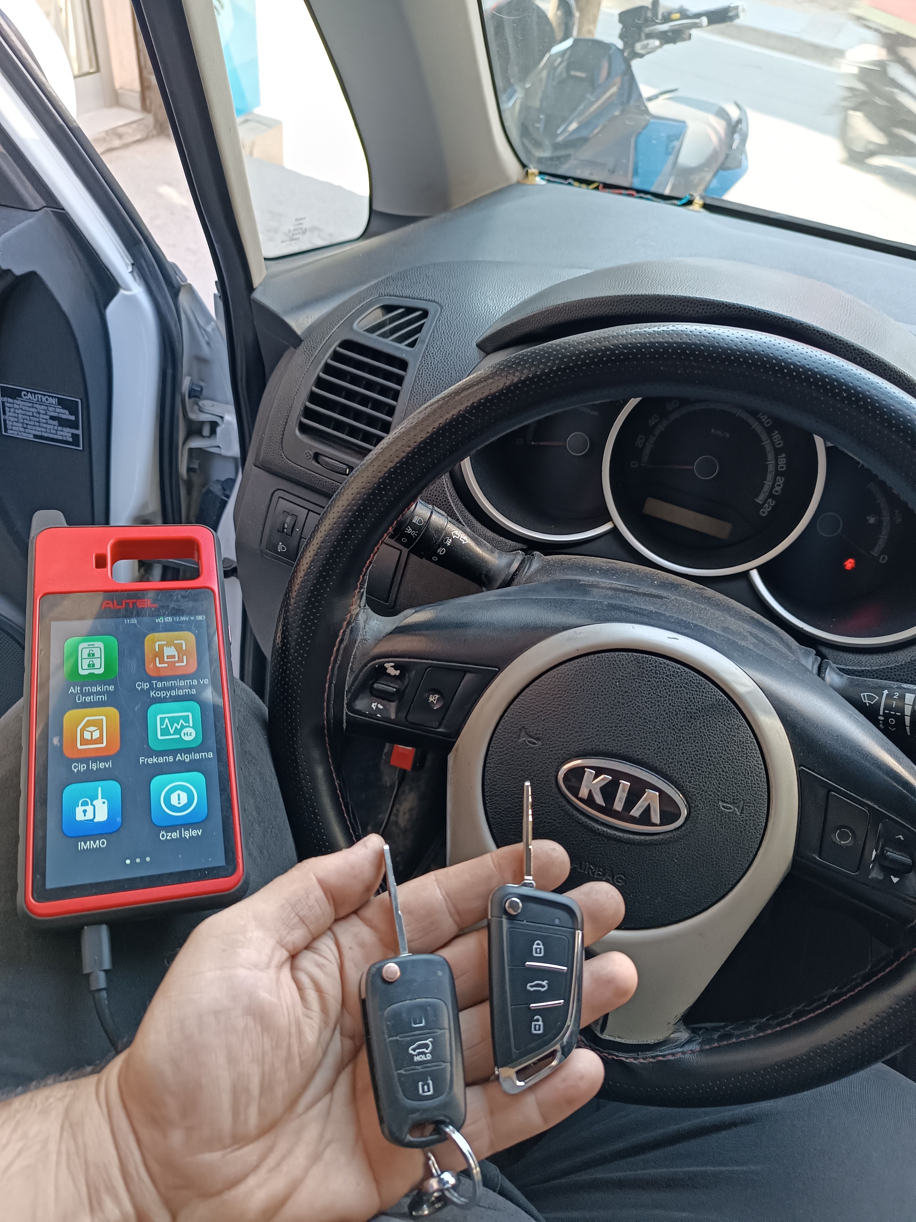 KIA direksiyonunun önünde programlama cihazı; elde eski ve yeni iki sustalı kumandalı anahtar